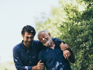 Jonge man wandelt met arm over de schouder van oude man, ze lachen en dragen allebei een blauw overhemd