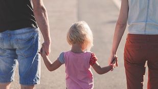 Closeup vanachter gezien van man in knielange spijkerbroek met zwart t-shirt en vrouw met oranje broek en witte bloes met tussen hen in, handen vast, een blond kind met roze t-shirt