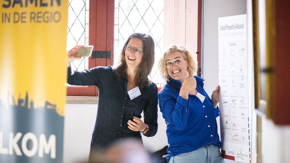 Vrouw met lang bruin haar in zwarte blouse maakt selfie met blonde vrouw in koningsblauwe blouse bij het bord over het Lokaal Preventie Akkoord