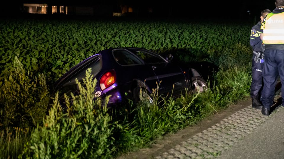 Donkerblauwe auto ligt op de zij in een sloot, twee agenten staan op de oever