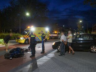 Midden op straat ligt een zwaarbeschadigde scooter met brokstukken er naast, erachter staat een ambulance en politie-auto, naast de scooter staan twee agenten en een paar omstanders
