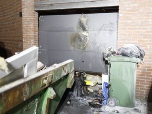Brandschade aan een roldeur van een garage met daarvoor verbrande rommel en op de voorgrond zie je nog net de hoek van een volle bouwafvalcontainer