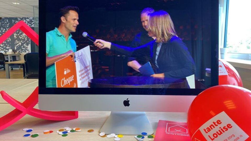 Rond computerscherm liggen ballonnen en confetti en op het scherm houdt een man in een turquoise shirt een oranje-witte cheque vast terwijl hij in microfoon spreekt van interviewer
