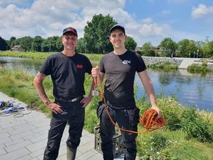 Twee mannen gekleed in zwarte broeken, zwarte t-shirts en basebalpetjes staan voor een brede sloot en één van hen houdt een oranje touw omhoog met een magneet er aan