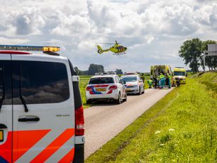 Op een smalle landweg staat een rij met politie-auto's met in de verte een ambulance waar hulpverleners bezig zijn rond een brancard en in de lucht komt een traumaheli aanvliegen