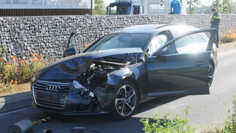 Donkere Audi met zwaarbeschadigde ingedeukte neus staat met beide deuren open midden op de weg en voor de auto liggen onderdelen van een andere auto, zoals een uitlaat