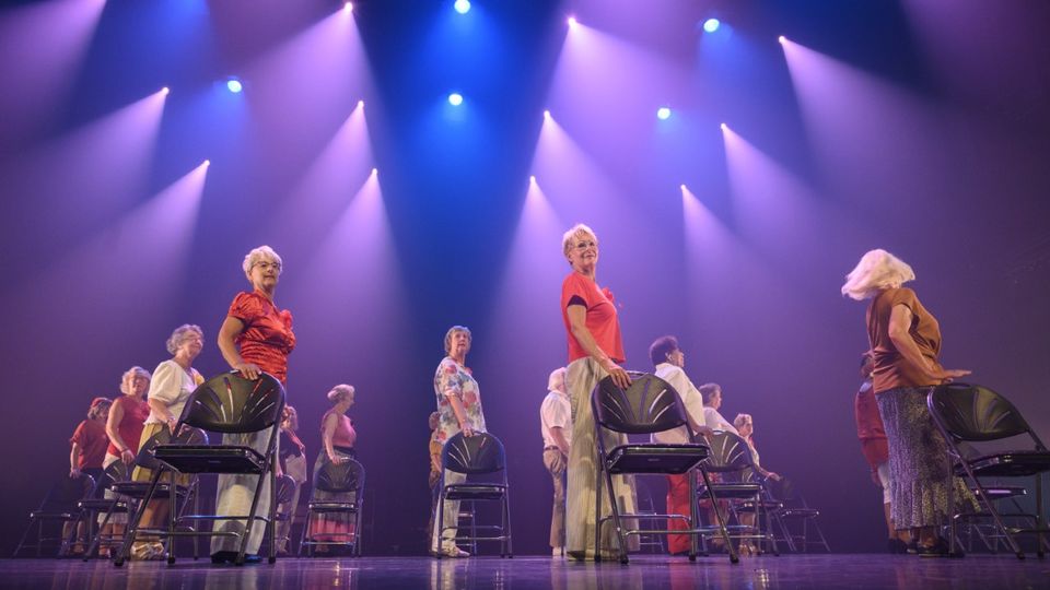 Paars uitgelicht podium met daarop een groep oudere dansers die ieder fier rechtop achter een stoel staan