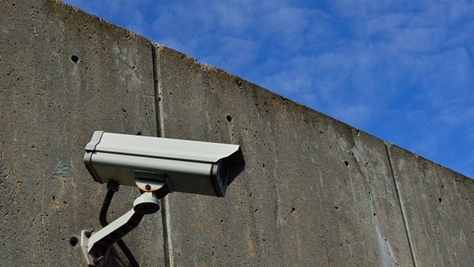 Beveiligingscamera gemonteerd op een betonnen muur met daarboven een lichtbewolkte blauwe lucht