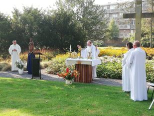Eucharistieviering in de open lucht met pastoor achter altaar naast het gazon met volwassen misdienaars aan weerszijden