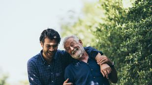 Jonge man wandelt met arm over de schouder van oude man, ze lachen en dragen allebei een blauw overhemd