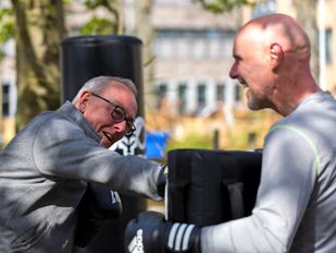Grijze man met bril in donkergrijze jas bokst tegen stootkussen dat vastgehouden wordt door een kale man in grijs shirt en zwarte bokshandschoenen
