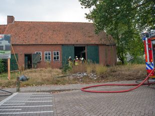 Op straat zie je nog net de achterkant van een brandweerauto met een uitgerolde rode brandslang en tussen de openstaande schuurdeuren van een oude boerderij met kapotte raampjes staan enkele brandweermensen