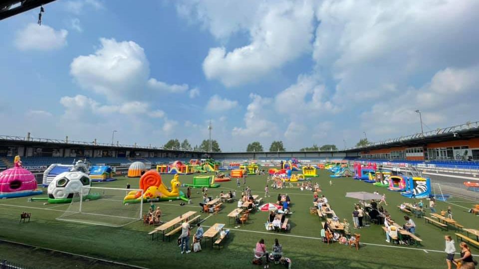 Zicht vanaf de stadiontribune op het voetbalveld dat vol staat met grote springkussens in allerlei vormen en kleuren en op de voorgrond zitten gezinnen aan picknicktafels