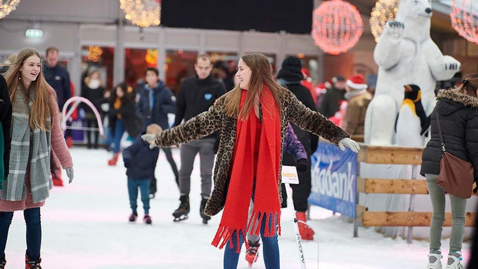 Schaatsers op ijsbaan met op de voorgrond een meisje in spijkerbroek, lange jas met dierenprint en knalrode sjaal dat schaatst met haar armen wijd open