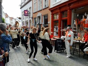 Publiek kijkt lachend toe als straatorkest en danseressen door smalle straat van Bergen op Zoom trekken