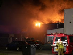 Brandweerman haalt spullen uit een brandweerbus terwijl op de achtergrond de vlammen uit het dak van een bedrijfspand slaan