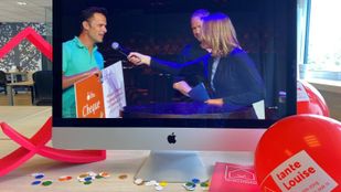 Rond computerscherm liggen ballonnen en confetti en op het scherm houdt een man in een turquoise shirt een oranje-witte cheque vast terwijl hij in microfoon spreekt van interviewer