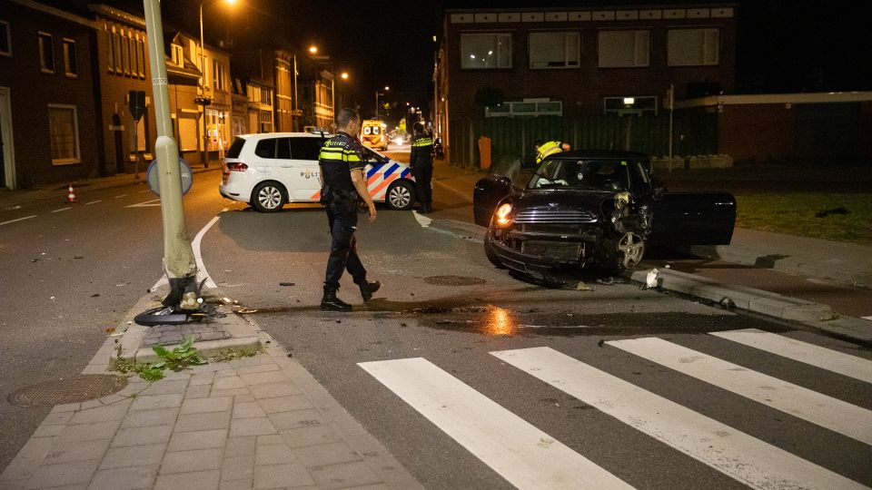 Een agent loopt langs een plas olie naar een zwarte auto die aan de voorkant volledig in elkaar zit en half op de stoep staat. In de middenberm staat een zwaar beschadigde lantaarnpaal.