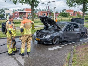 Twee brandweermensen in gele pakken staan bij zwarte uitgebrande auto waarvan de motorkap, deuren en kofferbak wijd open staan