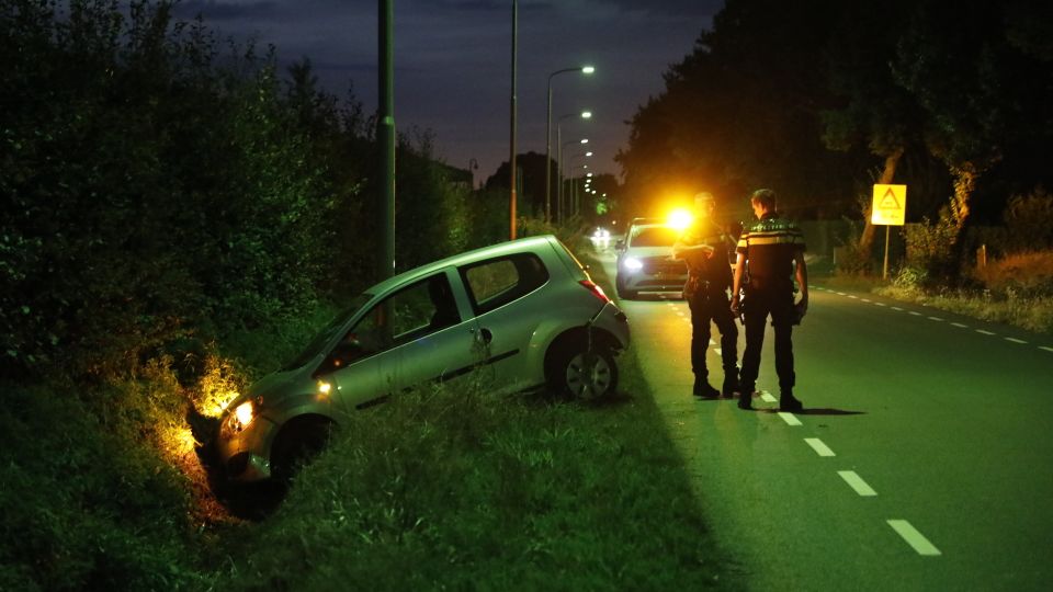 Licht gekleurde auto met hatchback hangt met de motorkap in een sloot, de koplampen branden nog en er staan twee agenten op de weg naast de beschadigde auto