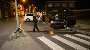 Een agent loopt langs een plas olie naar een zwarte auto die aan de voorkant volledig in elkaar zit en half op de stoep staat. In de middenberm staat een zwaar beschadigde lantaarnpaal.
