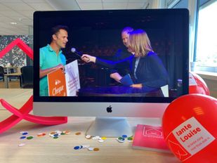 Rond computerscherm liggen ballonnen en confetti en op het scherm houdt een man in een turquoise shirt een oranje-witte cheque vast terwijl hij in microfoon spreekt van interviewer