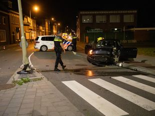 Een agent loopt langs een plas olie naar een zwarte auto die aan de voorkant volledig in elkaar zit en half op de stoep staat. In de middenberm staat een zwaar beschadigde lantaarnpaal.
