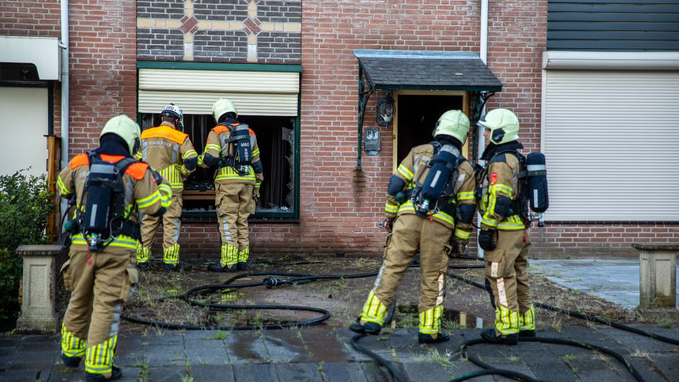 Vijf brandweermensen in de voortuin van een uitgebrand rijtjeshuis en twee van hen kijken via het kapotte raam naar binnen