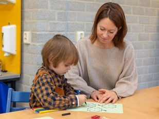 Vrouw kijkt mee met peuter die aan tafel zit te tekenen
