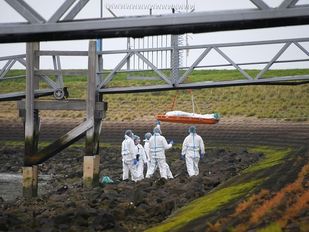Mannen in witte pakken staan bij afgedekt lichaam dat omhoog wordt getakeld