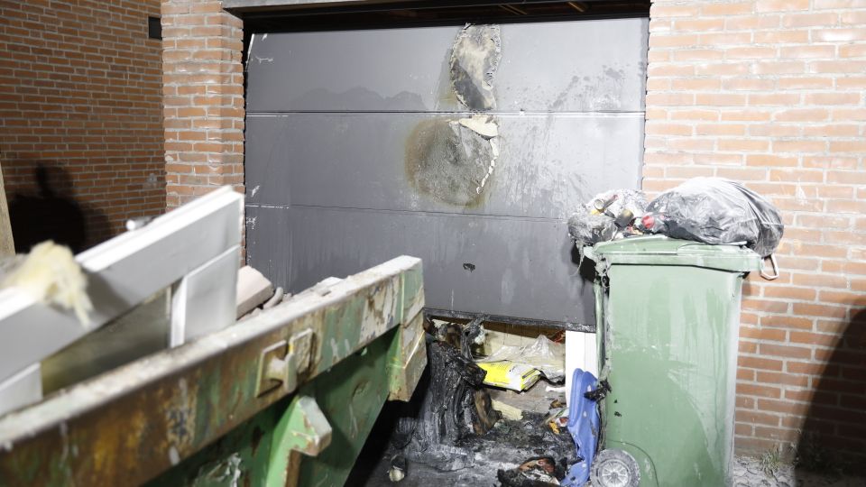 Brandschade aan een roldeur van een garage met daarvoor verbrande rommel en op de voorgrond zie je nog net de hoek van een volle bouwafvalcontainer