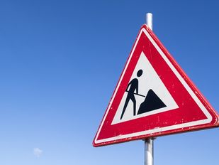 Onbewolkte helderblauwe lucht met op voorgrond recht een driehoekig verkeersbord wegwerkzaamheden met rode rand, witte achtergrond en zwart figuurtje dat zand schept.