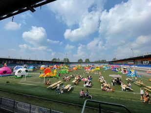 Zicht vanaf de stadiontribune op het voetbalveld dat vol staat met grote springkussens in allerlei vormen en kleuren en op de voorgrond zitten gezinnen aan picknicktafels