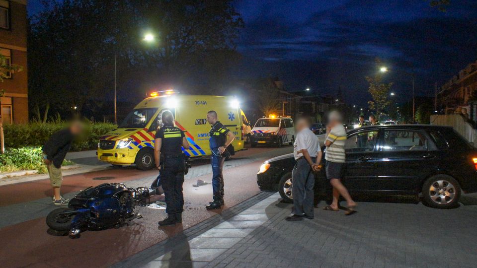 Midden op straat ligt een zwaarbeschadigde scooter met brokstukken er naast, erachter staat een ambulance en politie-auto, naast de scooter staan twee agenten en een paar omstanders