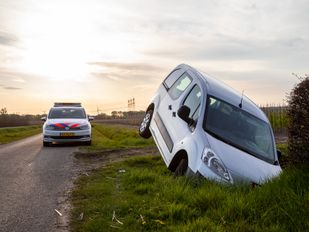 Zilverkleurige bestelwagen ligt met de neus vooruit en de achterbak omhoog in een sloot en op de achtergrond staat een politieauto