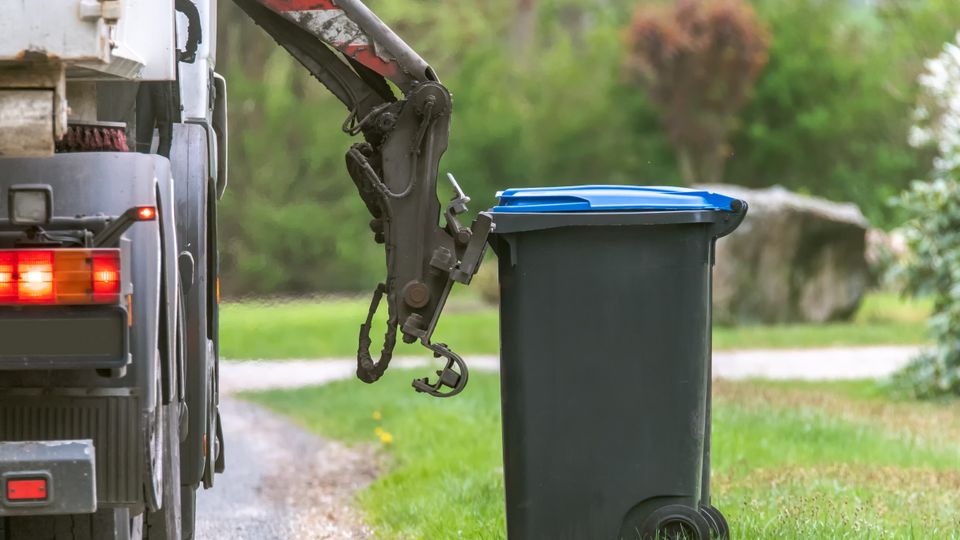 Grijparm van vuilniswagen koppelt zich aan een zwarte afvalcontainer met blauw deksel