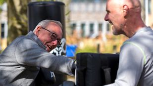 Grijze man met bril in donkergrijze jas bokst tegen stootkussen dat vastgehouden wordt door een kale man in grijs shirt en zwarte bokshandschoenen