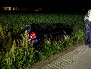 Donkerblauwe auto ligt op de zij in een sloot, twee agenten staan op de oever