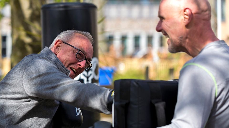 Grijze man met bril in donkergrijze jas bokst tegen stootkussen dat vastgehouden wordt door een kale man in grijs shirt en zwarte bokshandschoenen