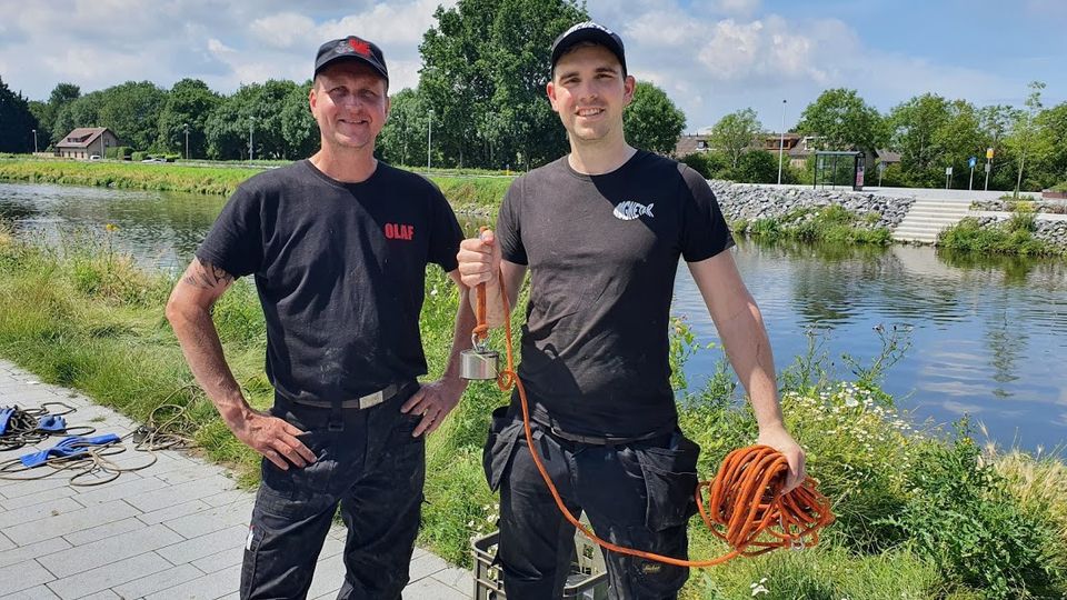 Twee mannen gekleed in zwarte broeken, zwarte t-shirts en basebalpetjes staan voor een brede sloot en één van hen houdt een oranje touw omhoog met een magneet er aan