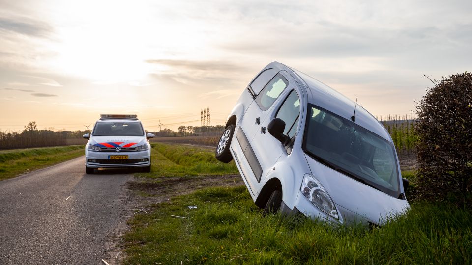 Zilverkleurige bestelwagen ligt met de neus vooruit en de achterbak omhoog in een sloot en op de achtergrond staat een politieauto