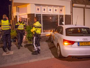 Drie agenten staan naast een witte auto die zich in de gevel van een hoekwoning heeft geboord.