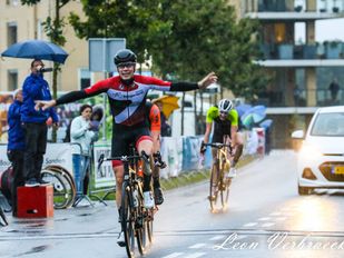 Wielrenner Remco Schouten rijdt in rood-wit-zwart shirt met armen wijd over finish in Hoogerheide langs publiek met paraplu's tegen de regen