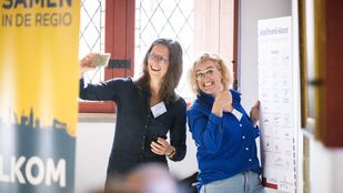 Vrouw met lang bruin haar in zwarte blouse maakt selfie met blonde vrouw in koningsblauwe blouse bij het bord over het Lokaal Preventie Akkoord
