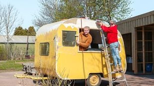 Gele caravan wordt geschuurd door vrijwilligers: eentje hangt uit het raam, eentje staat op een ladder tegen de buitenkant van de caravan