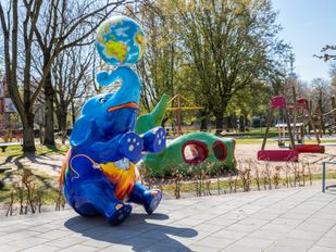 Kunstwerk van zittende blauwe olifant met geel-oranje clown op z'n buik en krullende slurf met daarop een wereldbol