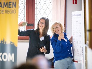 Vrouw met lang bruin haar in zwarte blouse maakt selfie met blonde vrouw in koningsblauwe blouse bij het bord over het Lokaal Preventie Akkoord