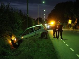 Licht gekleurde auto met hatchback hangt met de motorkap in een sloot, de koplampen branden nog en er staan twee agenten op de weg naast de beschadigde auto