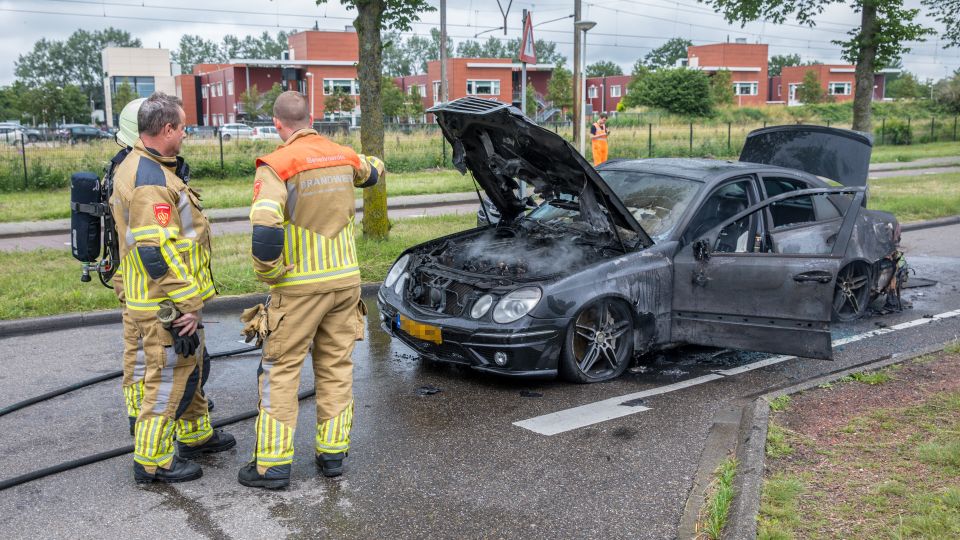 Twee brandweermensen in gele pakken staan bij zwarte uitgebrande auto waarvan de motorkap, deuren en kofferbak wijd open staan