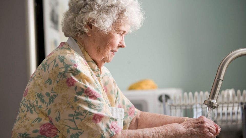 Oudere vrouw met kort grijs haar en bloemenbloes in pastelkleuren wast groenten onder de keukenkraan